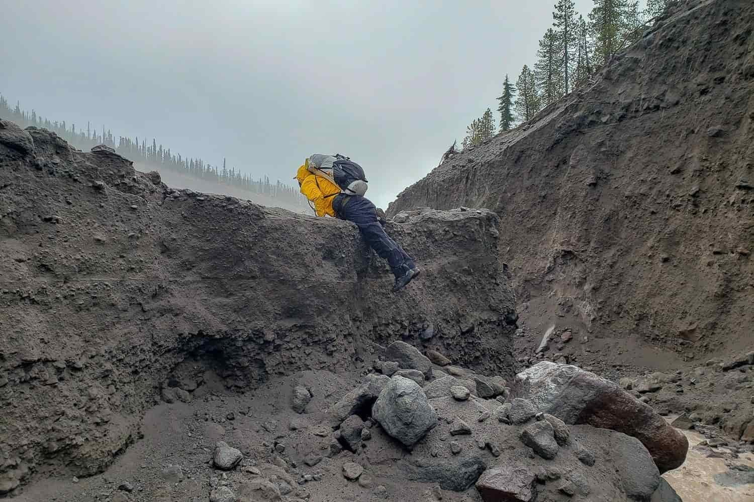 backpacker hoisting themselves up a washed out rugged river bank that is taller than the hiker.