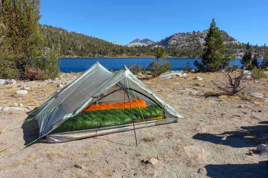 A tent set up next to a mountain lake with two sleeping bags visible through the tent's mesh