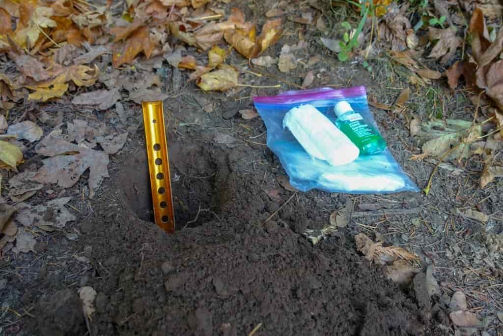 a cathole dug in the dirt with the trowel showing it is deep. there's hand sanitizer and toilet paper on the ground next to it