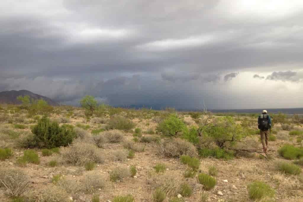 A hiker walks towards the dark gray horizon with a desert thunderstorm building
