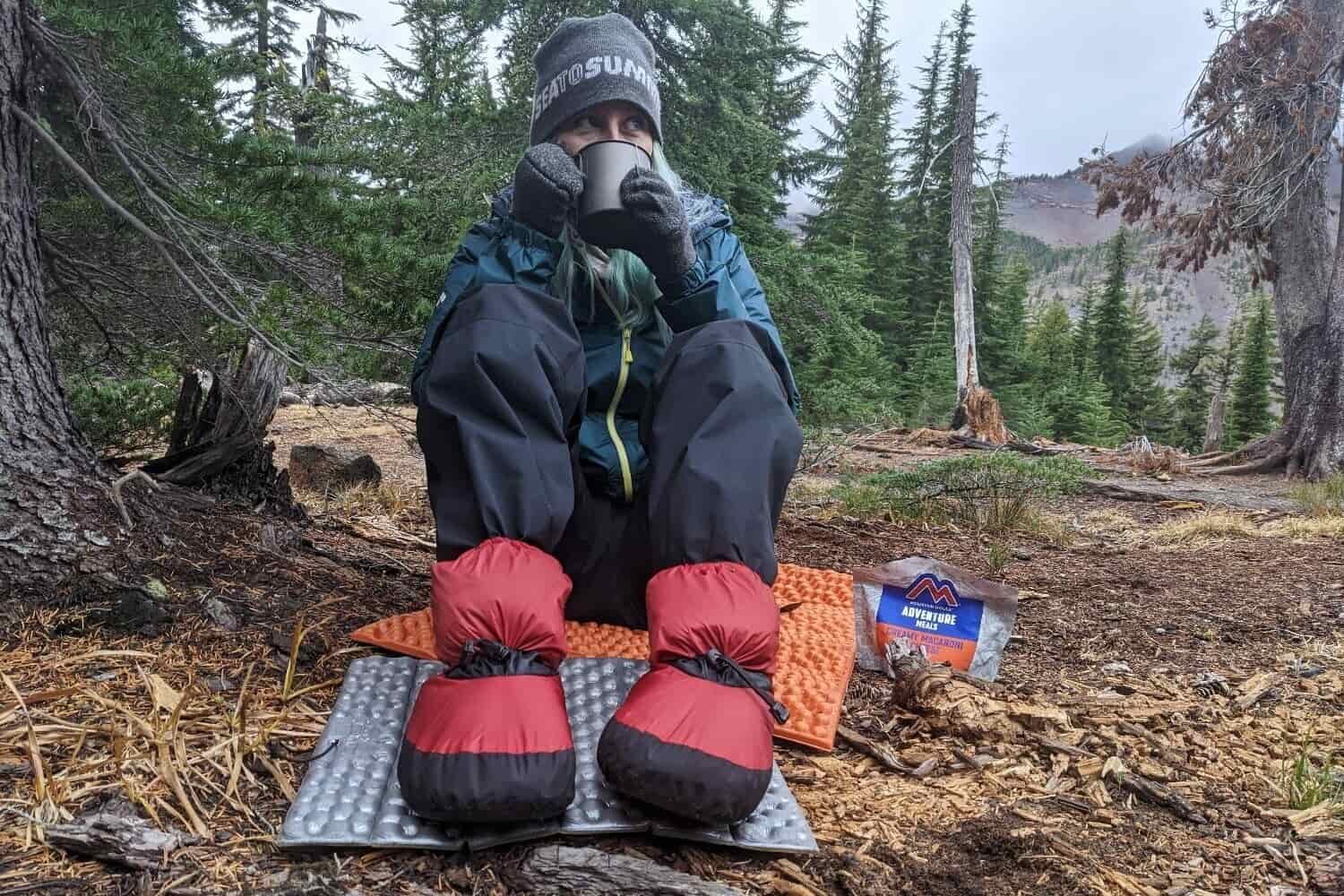 A-hiker-wearing-the-Feathered-Friends-Down-Booties-while-sipping-a-hot-drink-in-camp