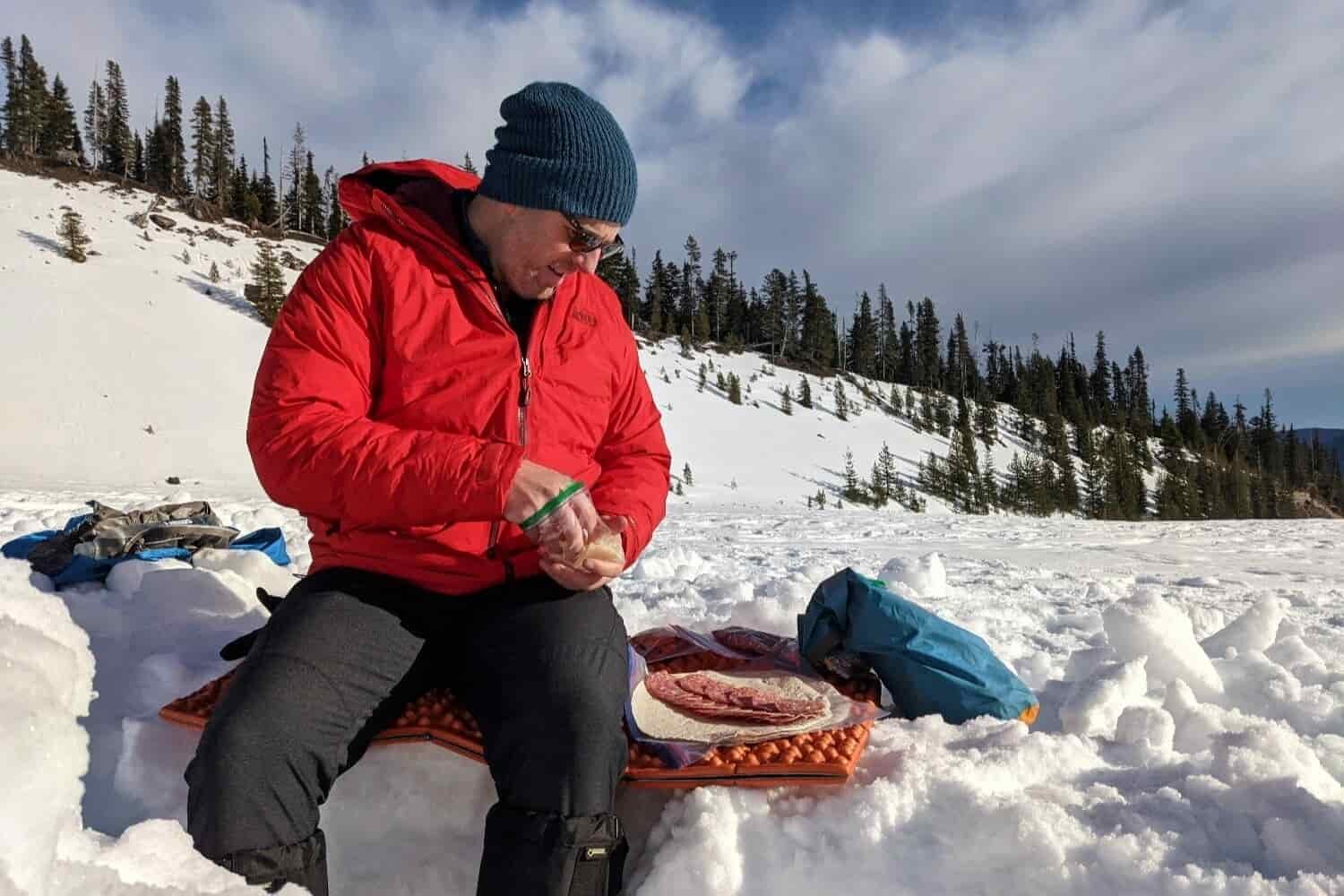 A-hiker-wearing-the-REI-Stormhenge-Jacket-while-eating-lunch-on-a-sunny-afternoon-during-a-winter-camping-trip