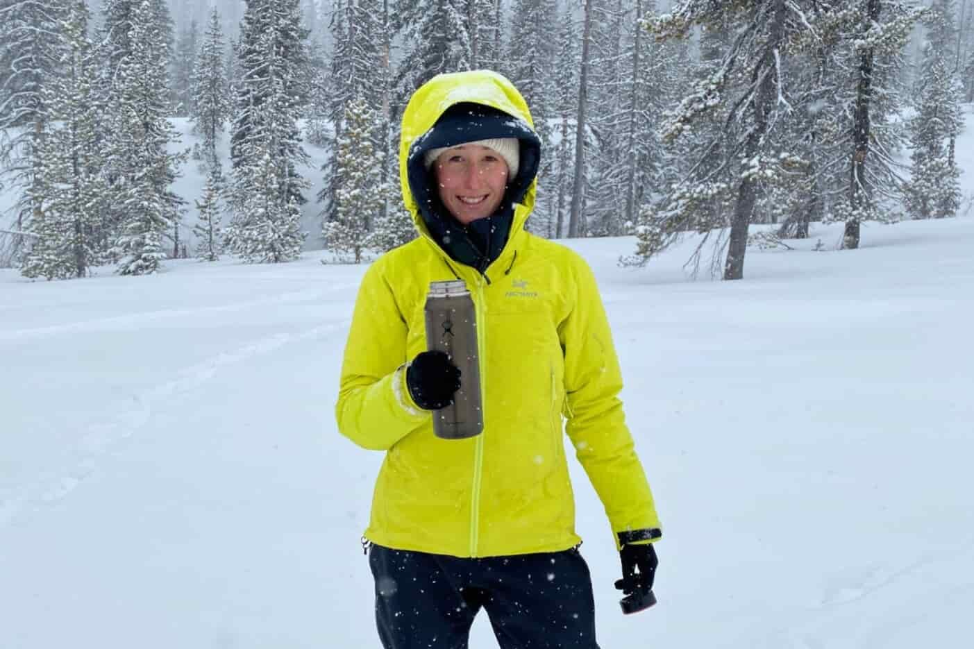 a women standing in a snowfield with snow covered firs in the background while holding a hot beverage in an insulated mug.