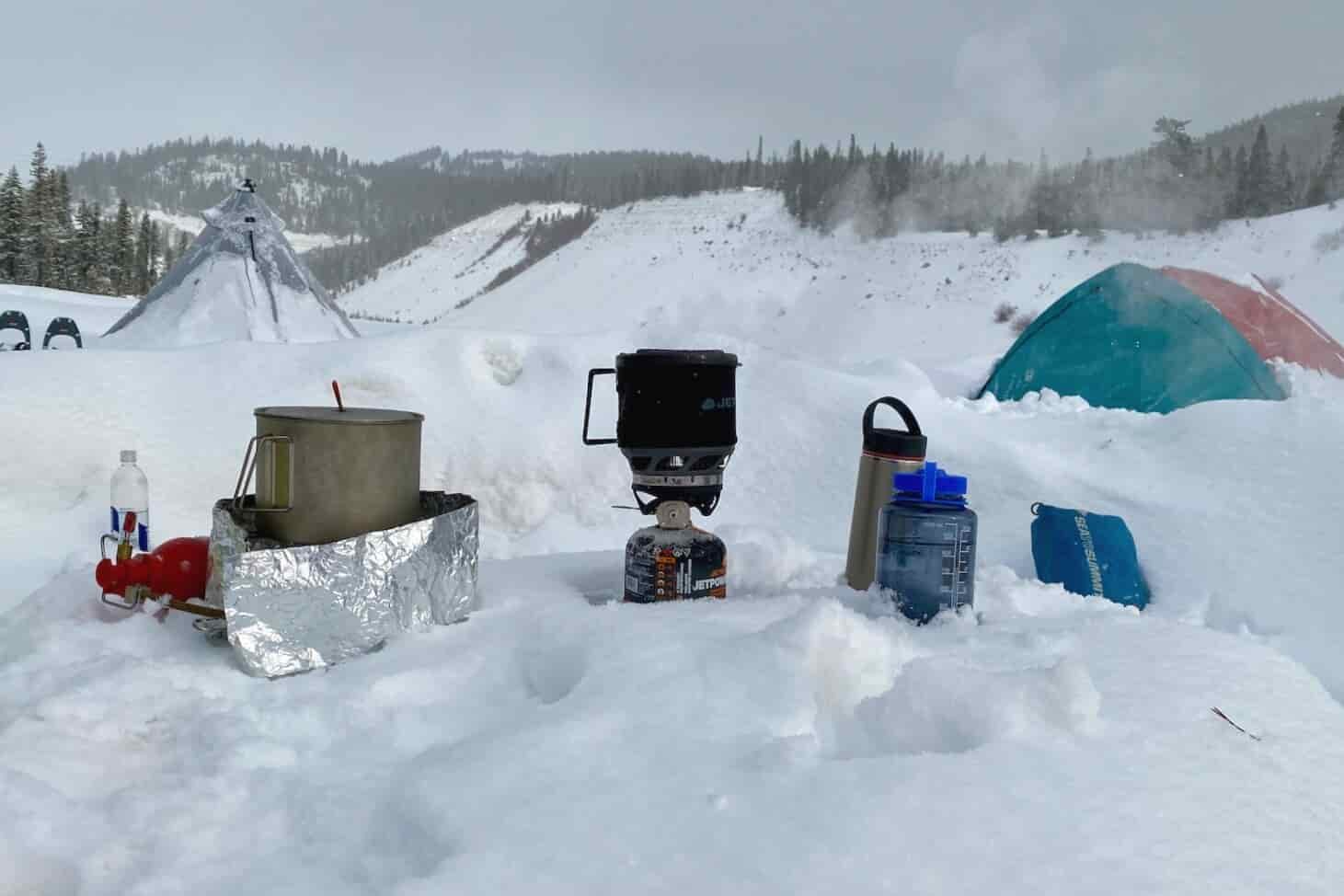 a kitchen on the snow with two camp stoves, water bottles and two snow covered tents in the background