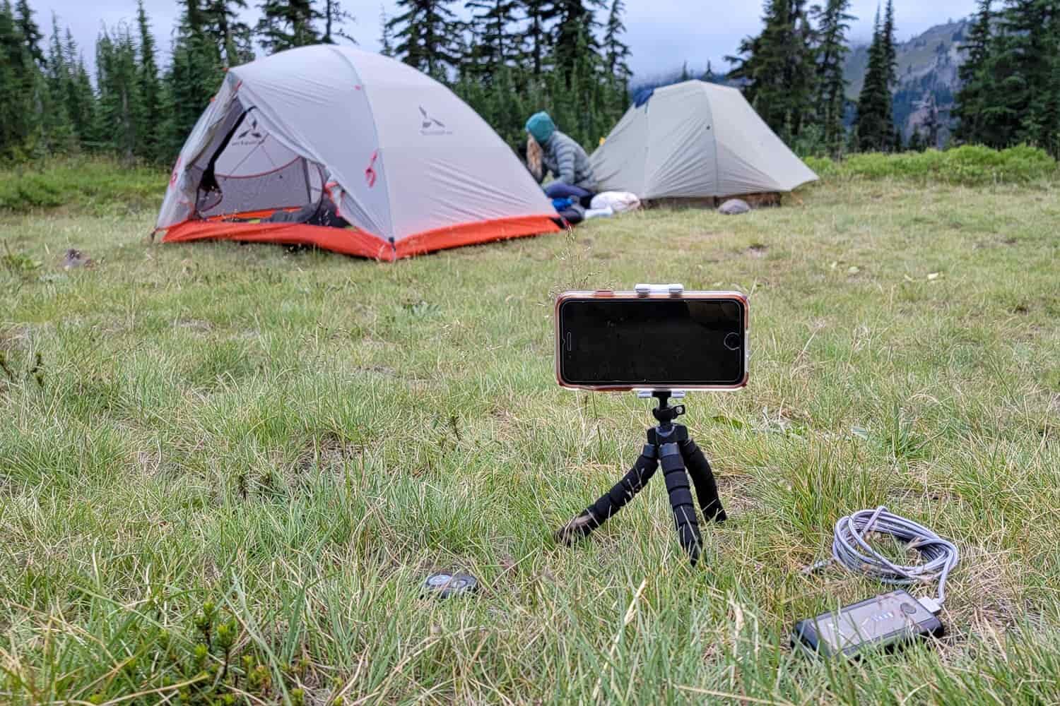 A cellphone sets on a tripod lining up a photography of a backpacker and two tents