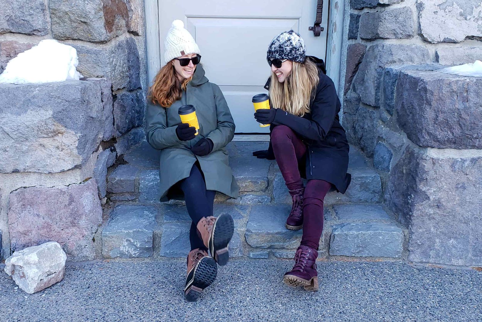 Two young women laugh while drinking coffee on a stone doorstep