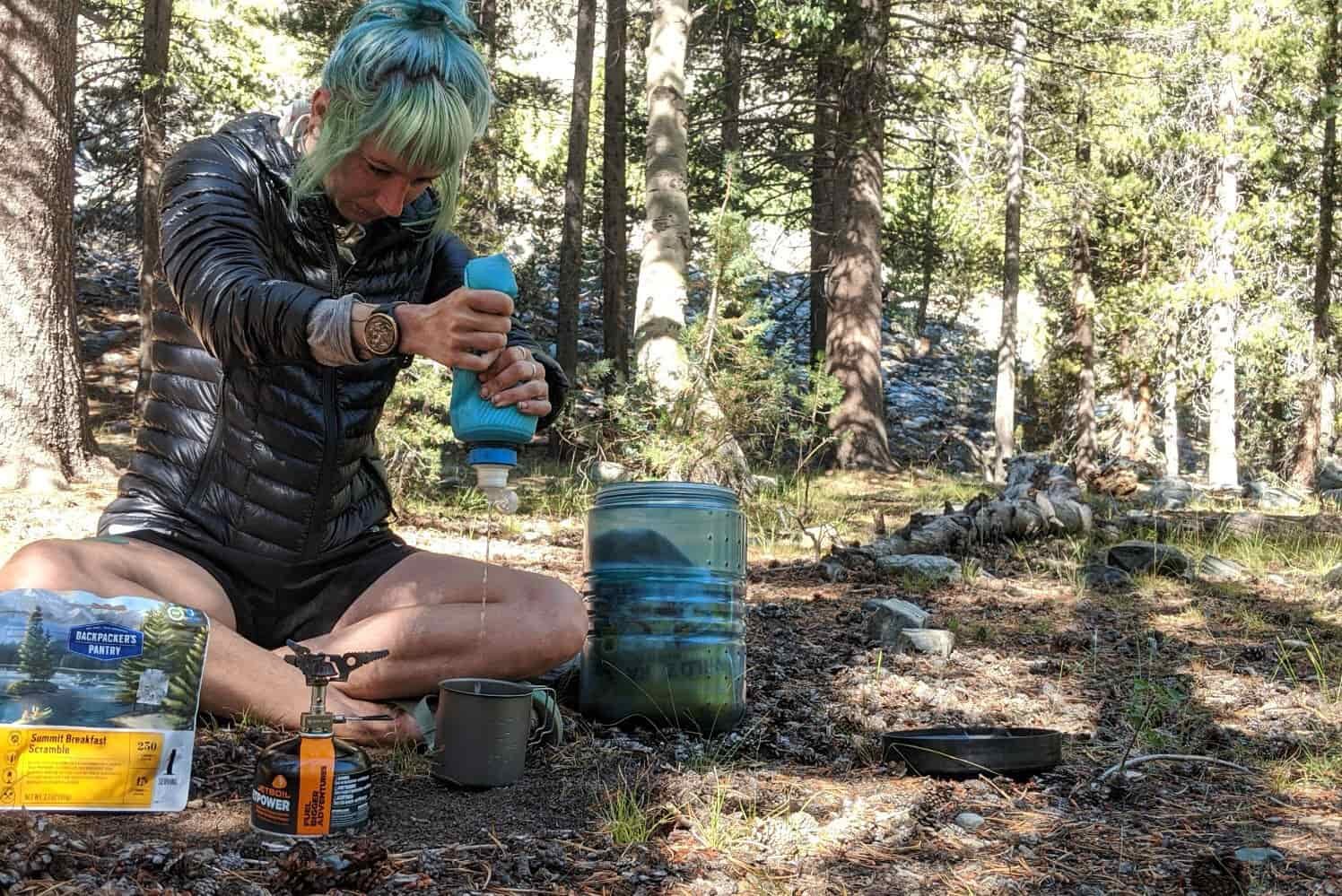 a hiker squeezes their water filtered water into a lightweight camping cup while making dinner in camp