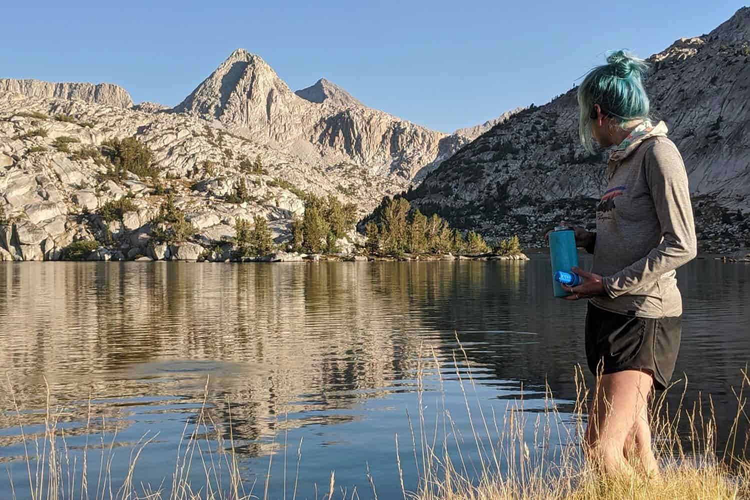 A backpacker reloading her water bottle and holding the filter that screws. She is in camp next to a lake surrounded by granite peaks.