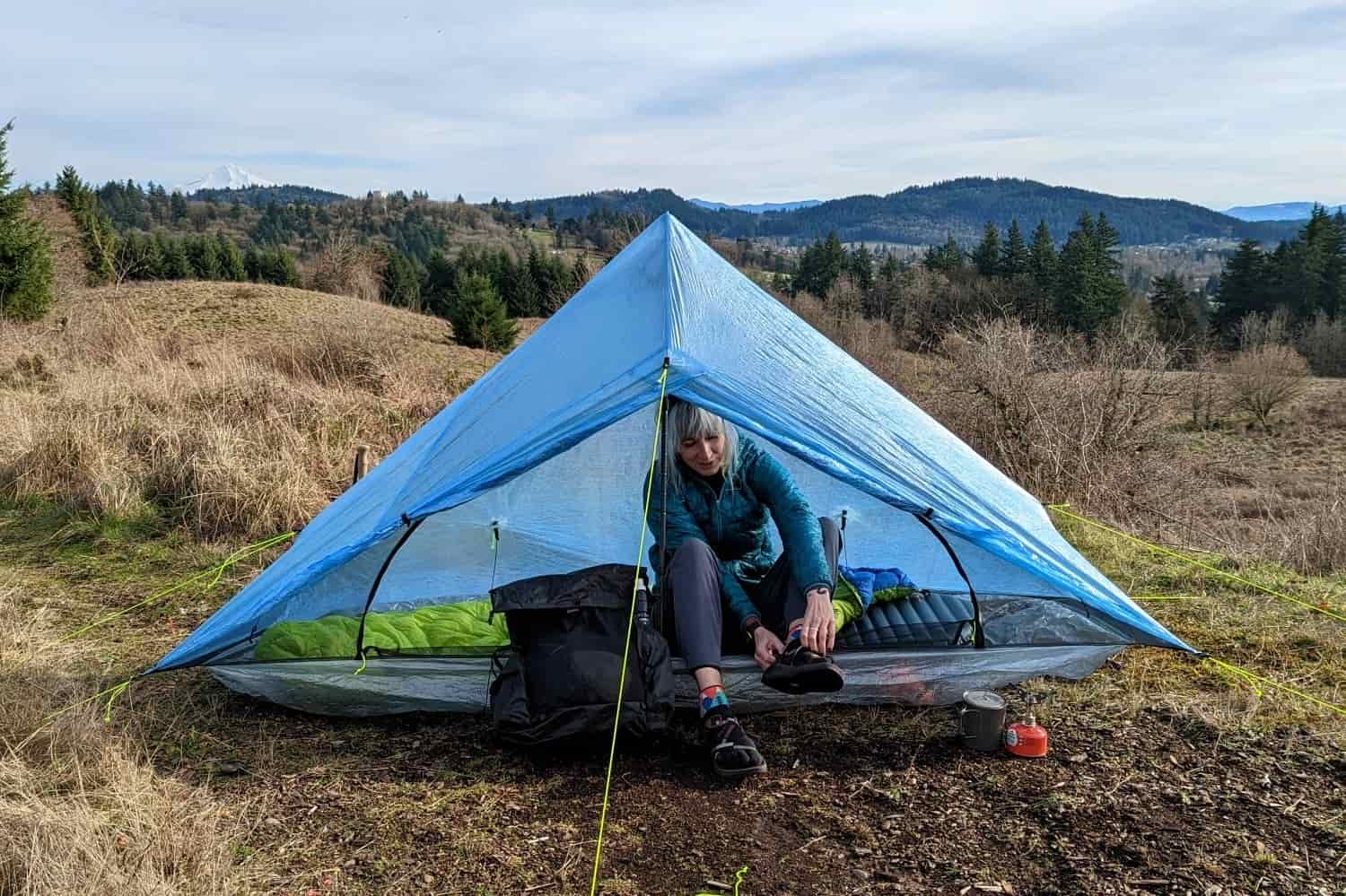 a backpacker sitting at the door of their zpacks plex solo tent putting their boots on with a snow covered volcano in the faint distance
