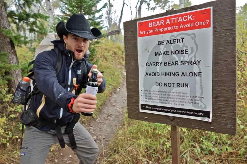 a backpacker holding up bear spray to the camera next to a sign that informs hikers how to avoid a bear attack.
