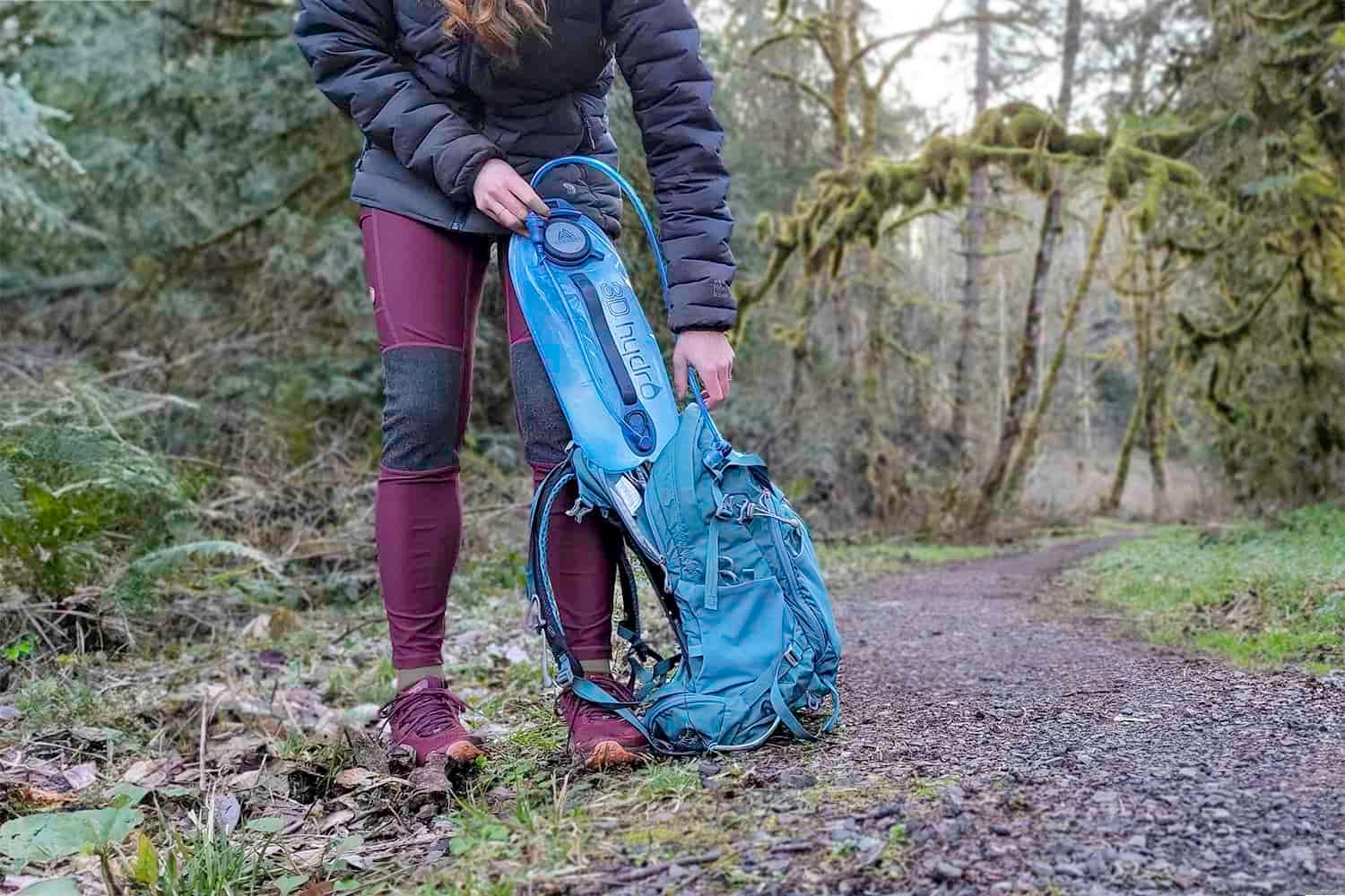 Waist-down view of a female hiker pulling the Gregory 3D Hydro hydration bladder out of her backpack along a trail