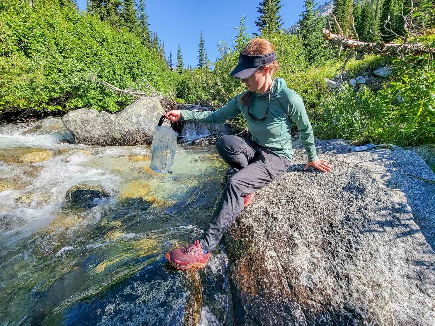 backpacker on a boulder near a clear river filling up the dirty water resevoir to treat the water for safe drinking