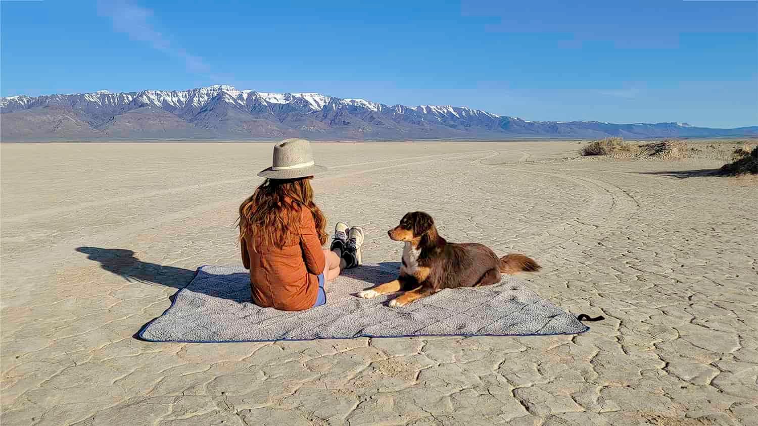A woman and a dog sitting on the Oceas Sherpa Fleece camping blanket in the Alvord Desert with the Steens Mountains in the background