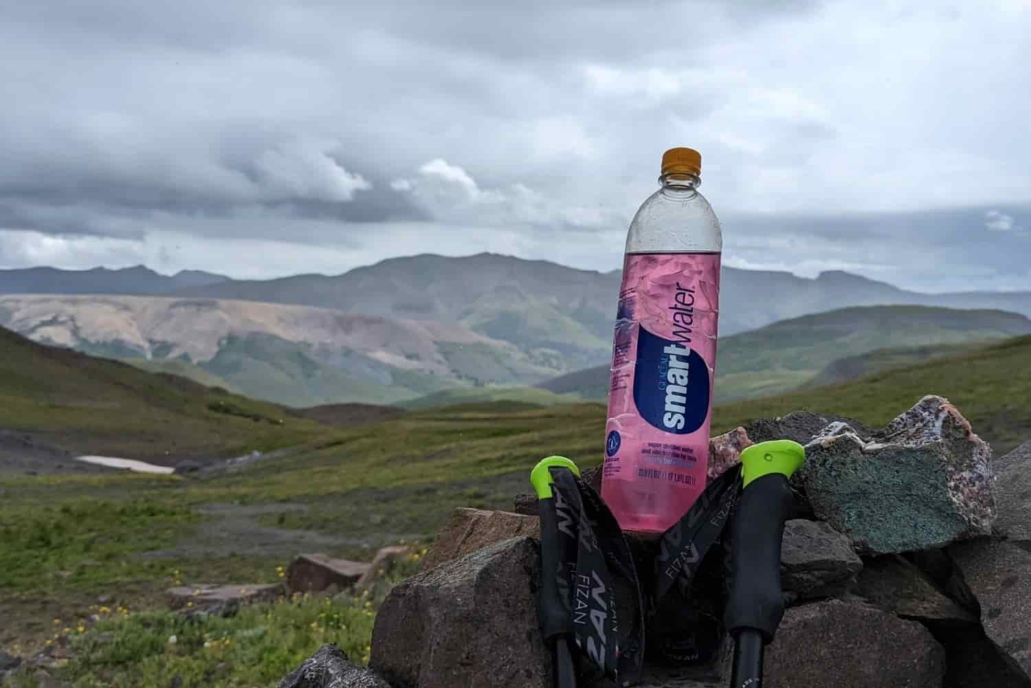 a smart water bottle with a water treatment tablet and an electrolyte mix with a view of sloping hills and clouds in the background and trekking poles propped next to the water bottle