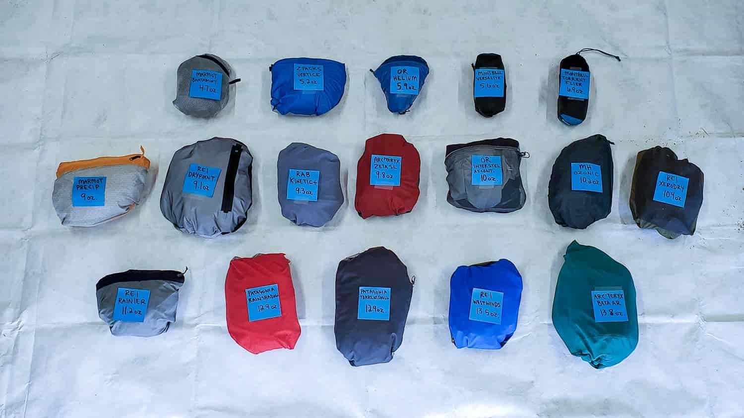 13 rain jackets stuffed into their sack or stash pocket with their brand and weight labeled to compare the weight and packability of each option