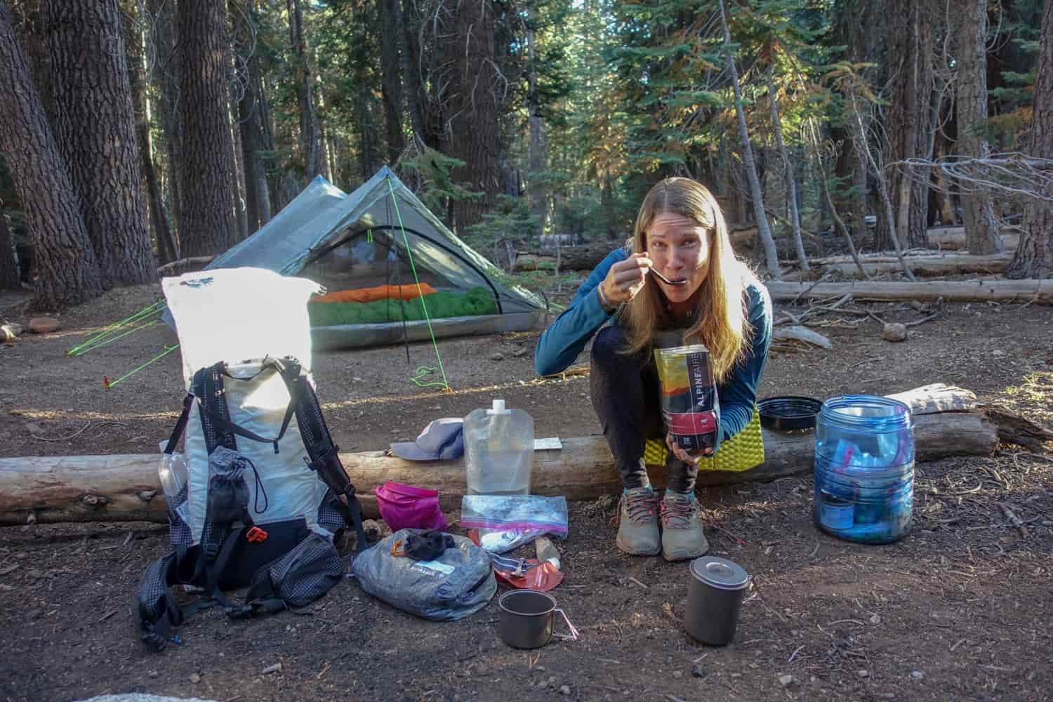 backpacker on the JMT in camp with a tent set up and her backpack emptied while eating a dehydrated meal.