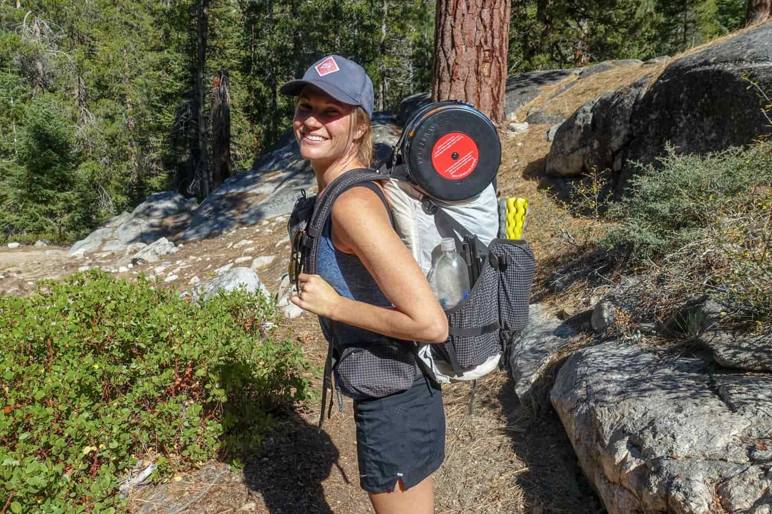 Backpacker showing her fully loaded pack with a pad, water bottle, bear canister, and tent poles all strapped to her backpack.