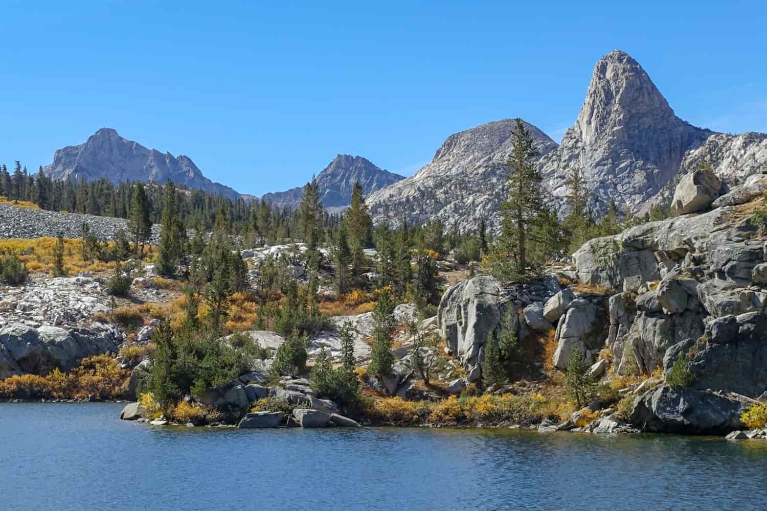 Granite spires behind a boulder field with evergreen trees and golden brush with a lake in the foreground.