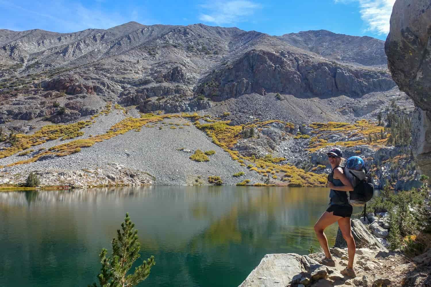 Backpack poses for a a photo on the trail along a lake's edge with a rock wall overhanging the trail.