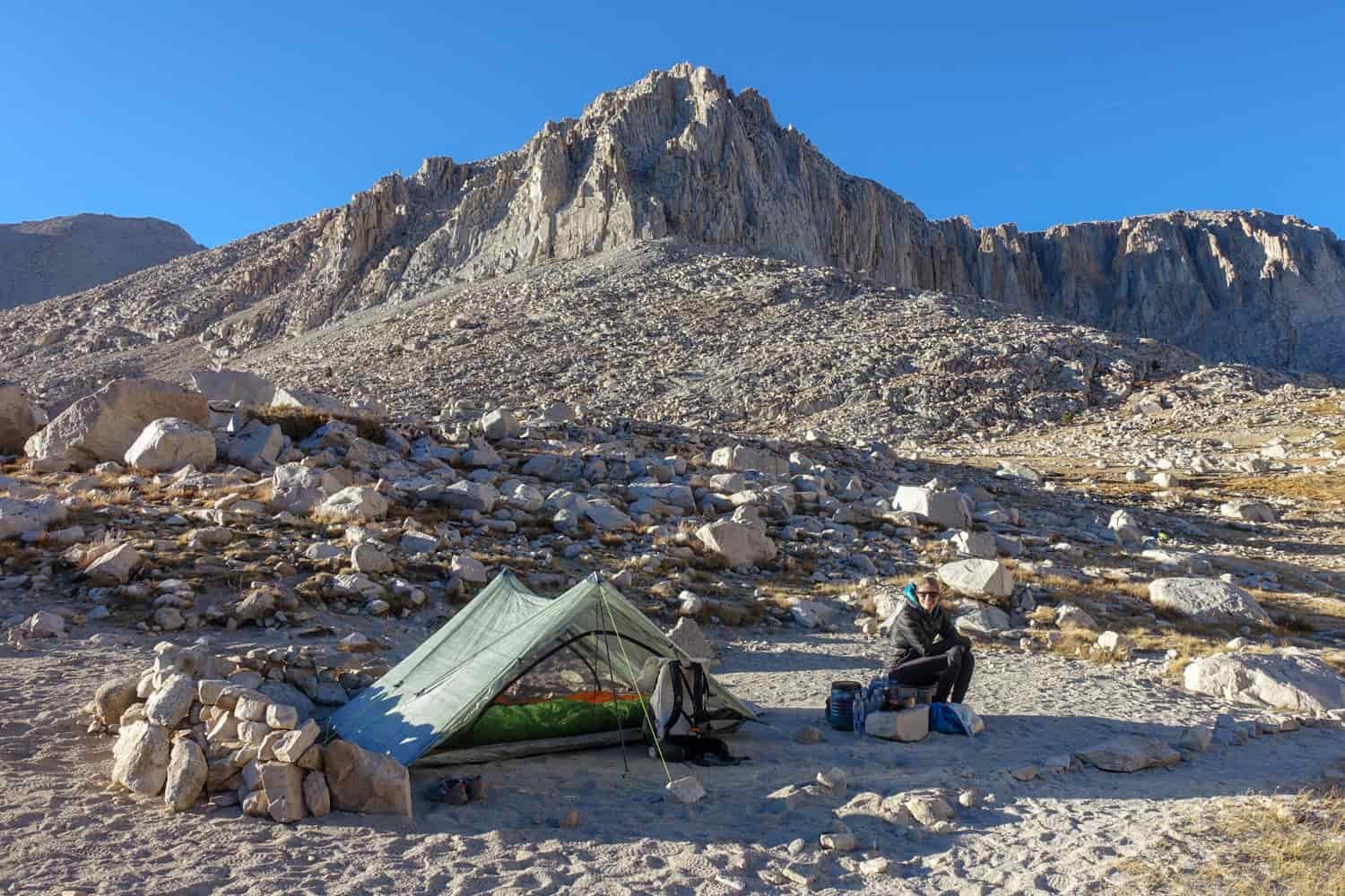 Tent pitched in a rocky alpine tundra with a backpacker sitting beside tent.