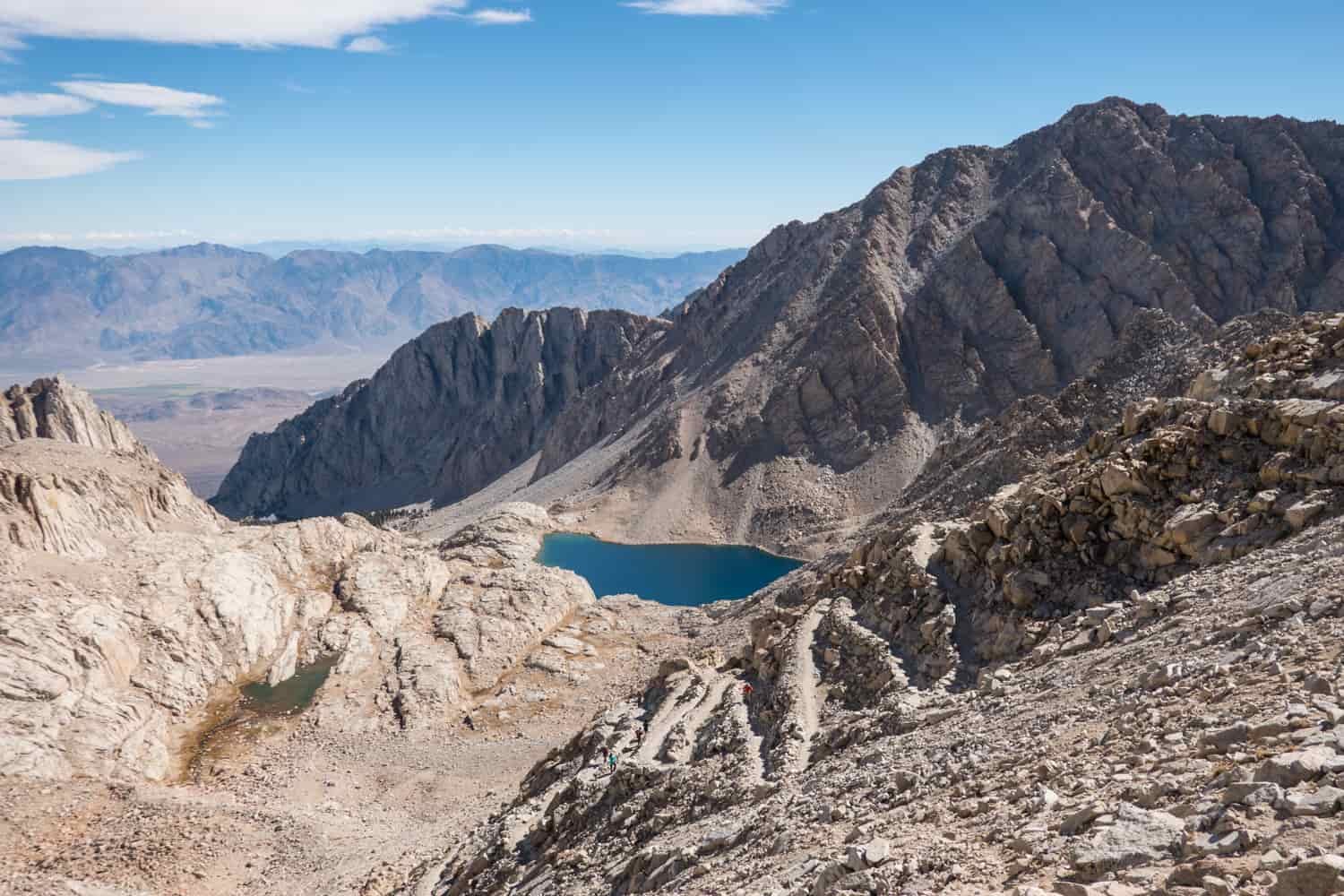 A blue tarn surrounded by mountains with the valley floor in the background. A well made trail with several switchbacks and a backpacker hiking down them in the the foreground.
