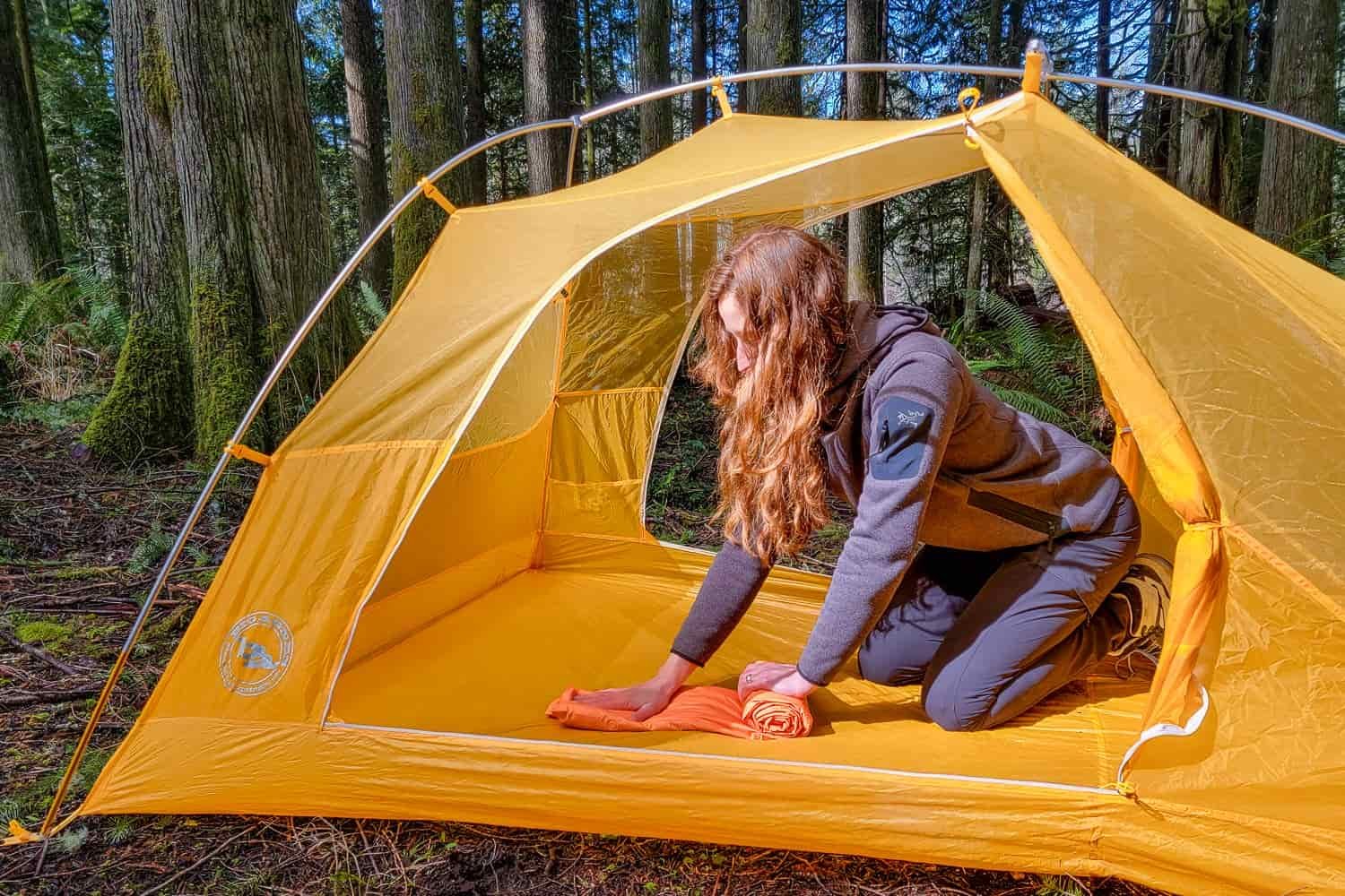 a backpacker rolls the Big Agnes Zoom Ultralight sleeping pad inside a tent with the door rolled open in a forest.