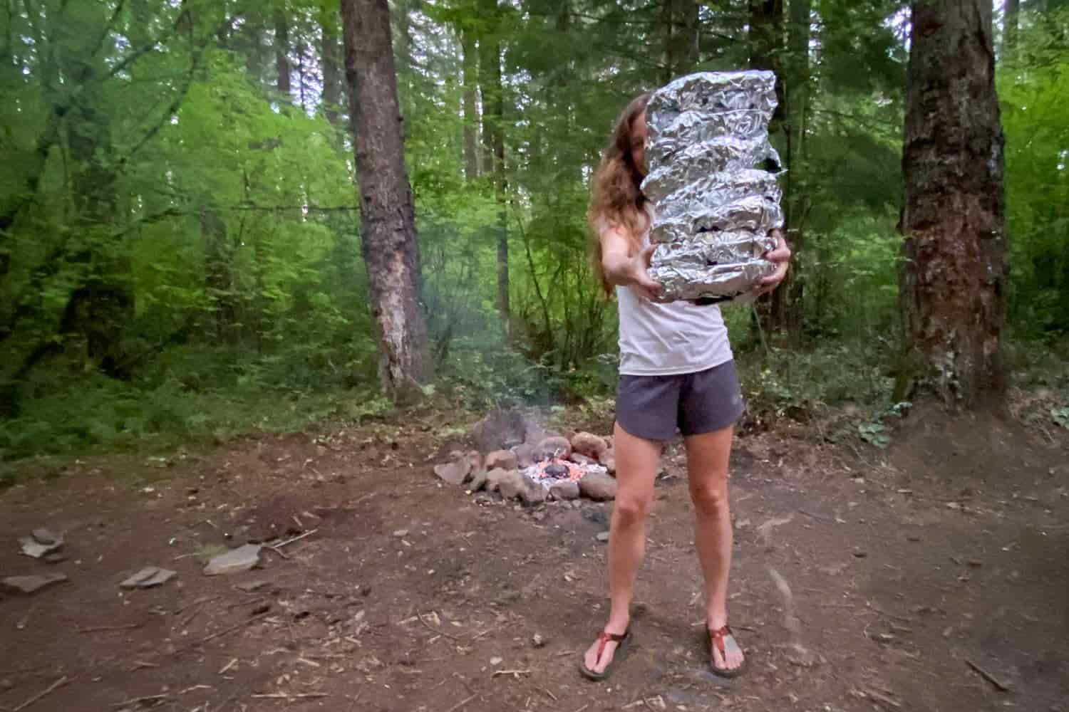 person in a campsite holding meals wrapped in foil