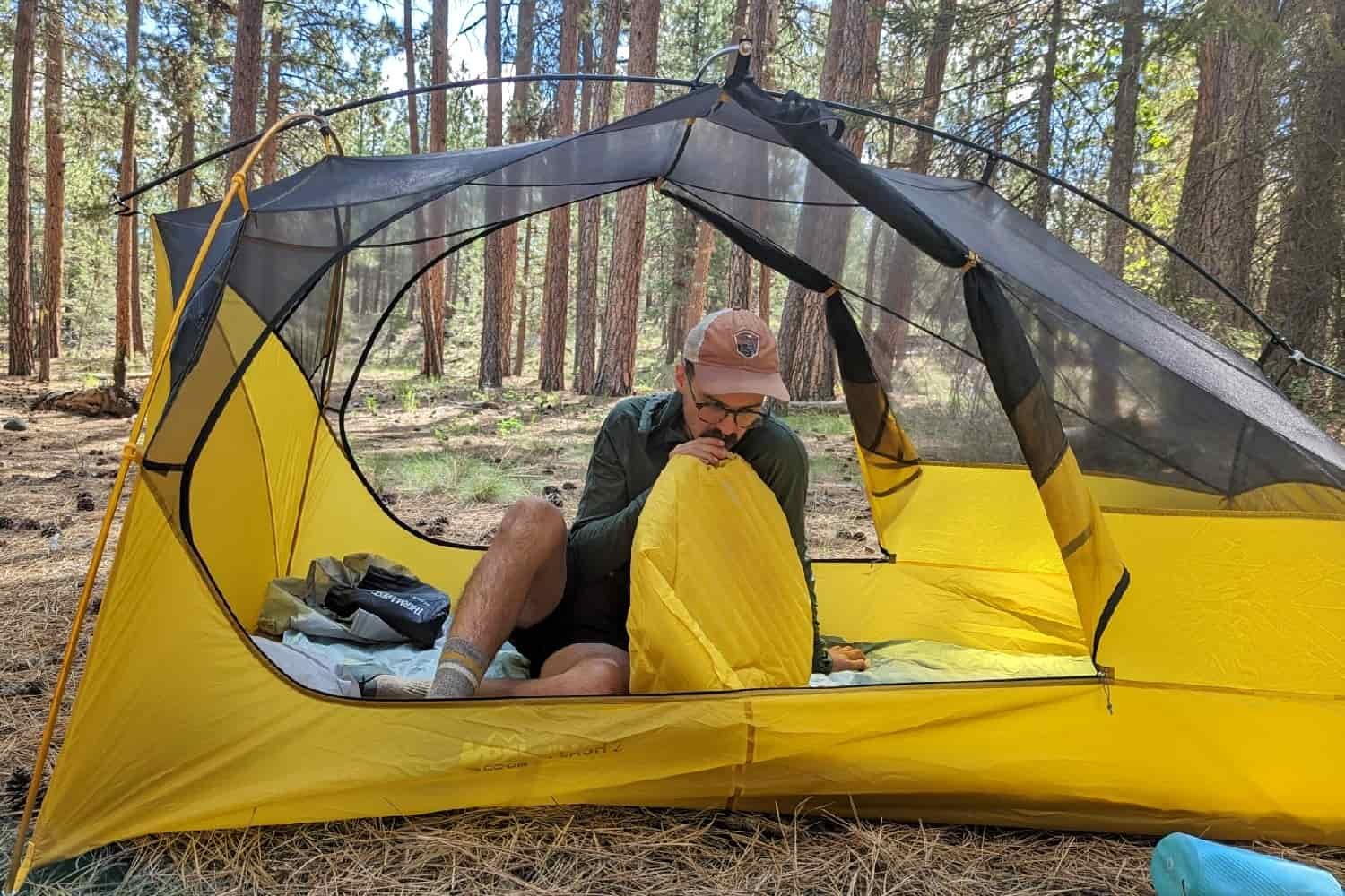 a backpacker sitting inside their open tent blowing up their sleeping pad with their mouth