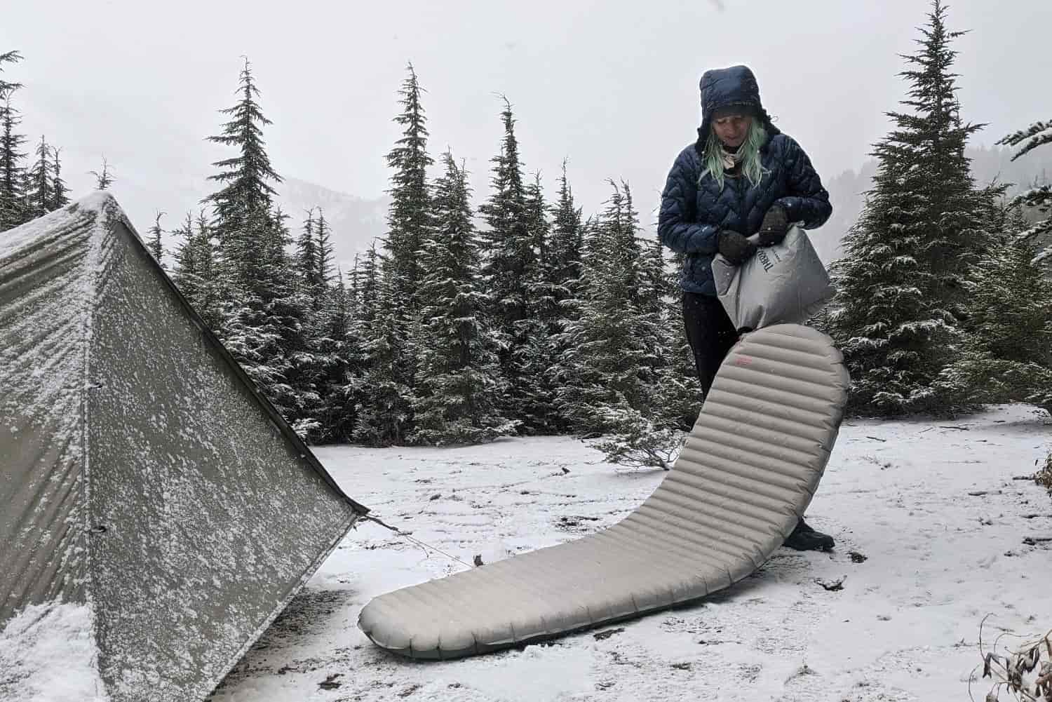 a hiker in a snow covered meadow inflates their sleeping pad while standing next to their tent that is covered in snow.