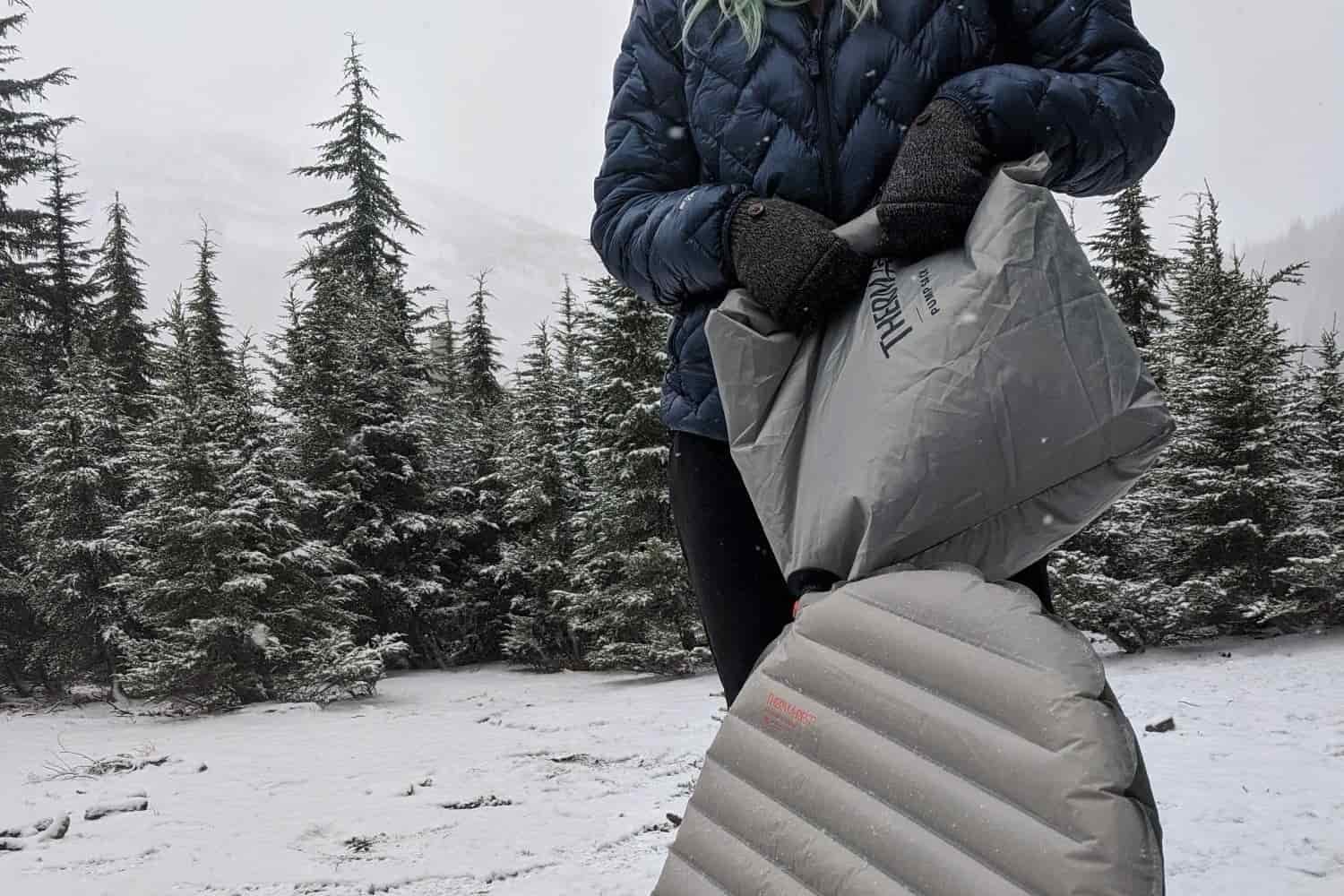 a snow covered ground and firs in the background while a thru-hiker wearing jackets and gloves inflates their NeoAir Xtherm NXT with the pump sack.