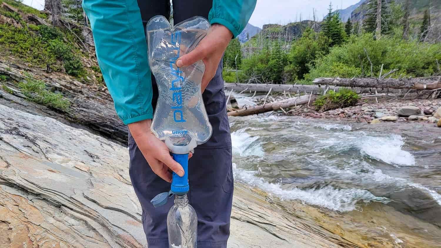 a backpacker stands near a stream with a water bottle on the ground between their feet for support while they squeeze a quickdraw water bladder through a filter.