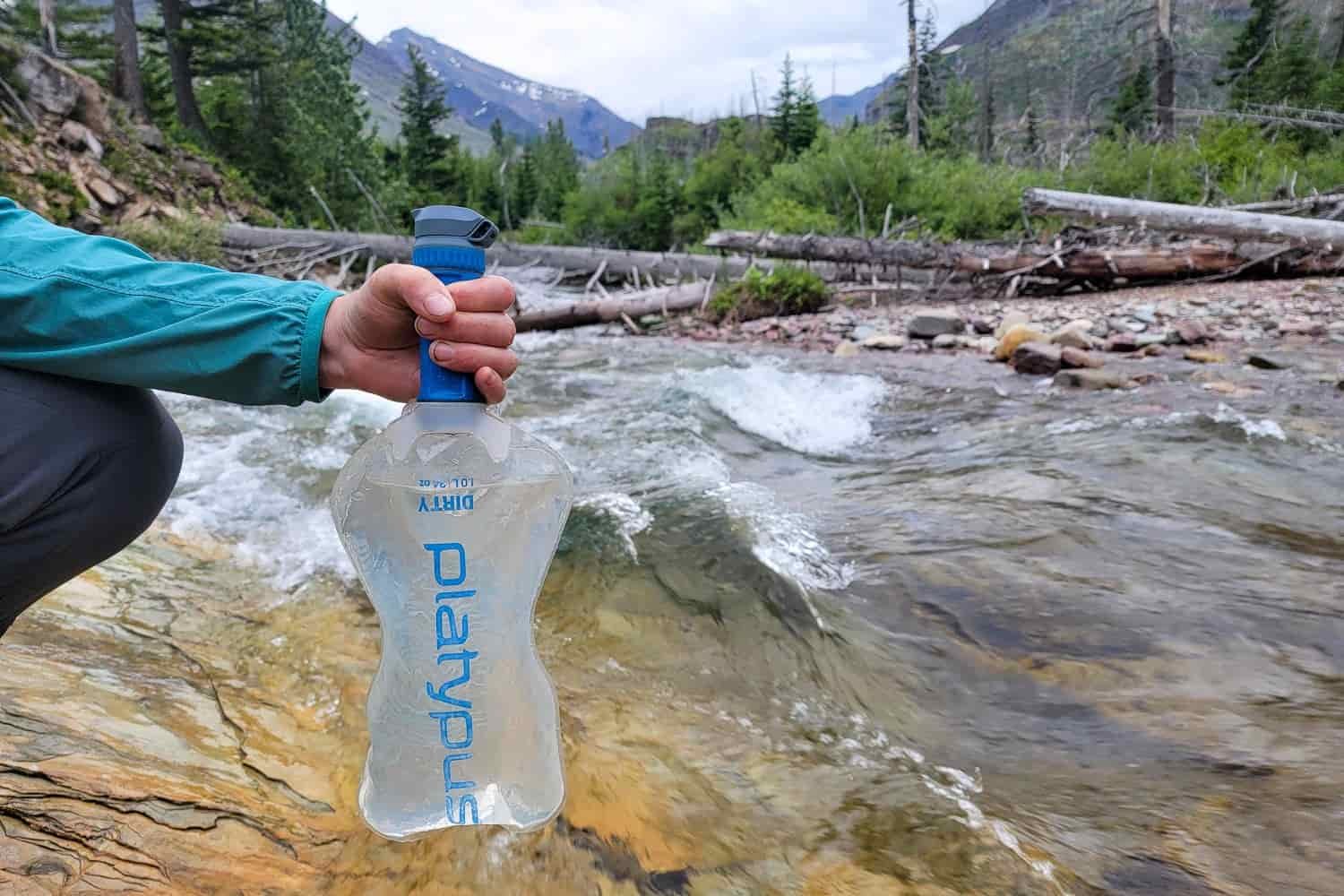 Closeup of a hiker gripping the filter cartridge of the Platypus QuickDraw with a rushing river behind and mountains in the distance.