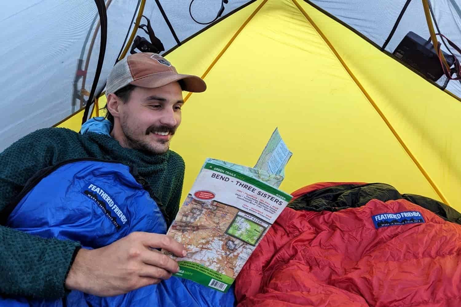 A man reads inside a blue sleeping bag inside a tent while backpacking.
