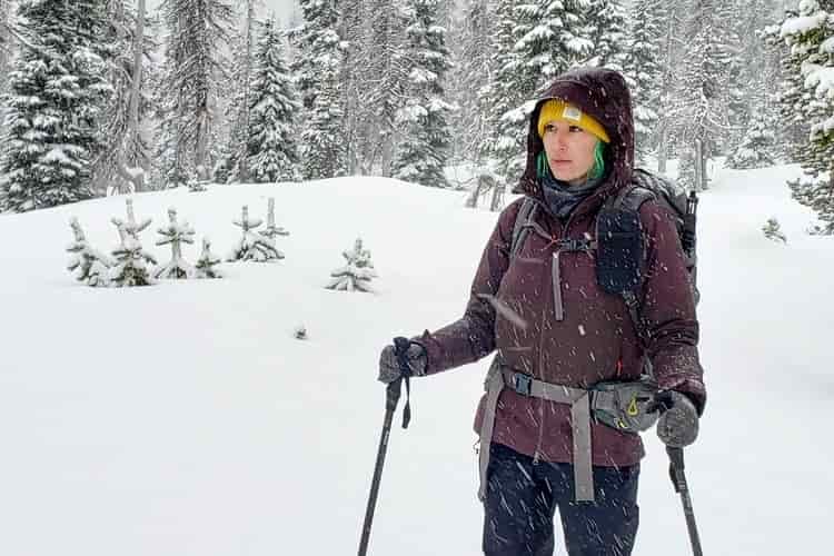 A female backpacker stands in a snow flurry wearing a purple insulated jacket and yellow beanie