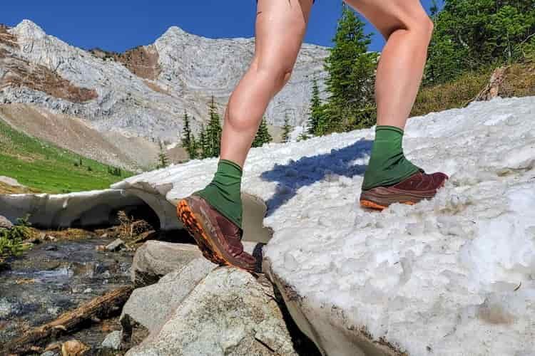 A female hiker steps from a rock onto a snowbank in a pair of trail running shoes with gaiters over her ankles 