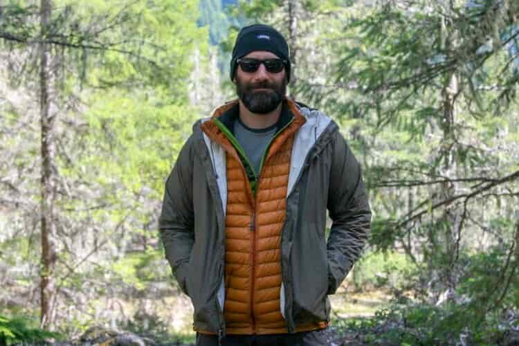 A bearded hiker layered up with a beanie on his head, sunglasses on his face, and two puffy jackets and a rain shell layered over a baselayer