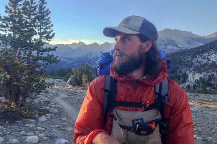 A bearded backpacker looks to his right while wearing a cap and windbreaker jacket layered over an insulated midlayer