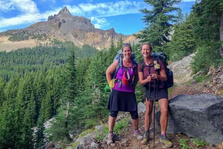 Two female hikers smile on a mountain trail wearing light synthetic hiking tops and skorts on the bottom
