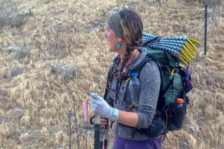 An ultralight backpacker with a buff on her head, lightweight fleece top on her torso, and small backpack topped with a sleeping pad