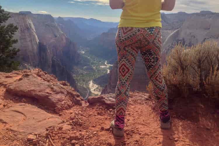 Rear view of a female hiker in colorful leggings as she stands on a red rock cliff looking down on a green valley