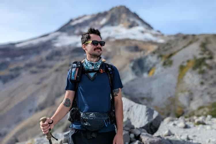 A male hiker in a blue synthetic t-shirt is in focus with a mountain out of focus in the background
