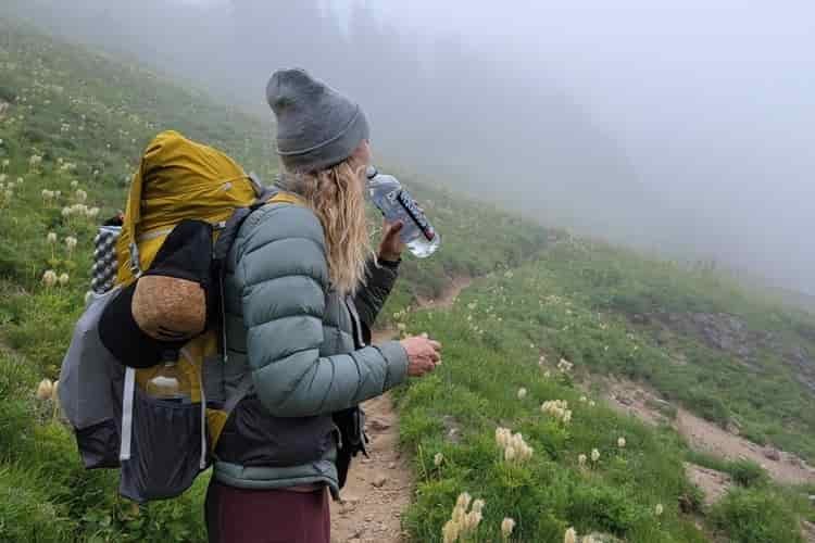 A female backpacker sips water from a water bottle on a foggy trail while wearing an insulated jacket, beanie, and backpacking pack