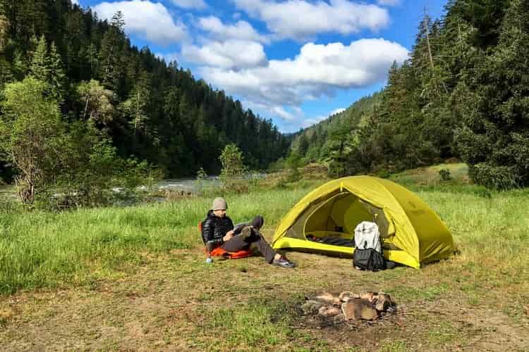 A male hiker reclines next to a yellow tent while reading 