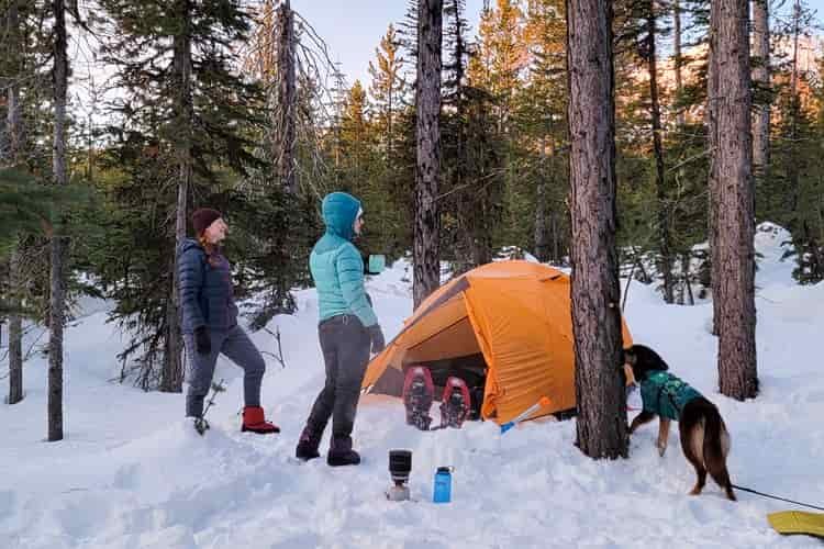 Two female backpackers and a dog stand next to a tent pitched in the snow while wearing down hooded jackets, down pants, and down booties to stay warm