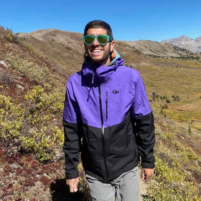 Against a summer mountain backdrop in the Colorado Rockies, a man in purple and black rain jacket and green sunglasses hikes towards camera while smiling