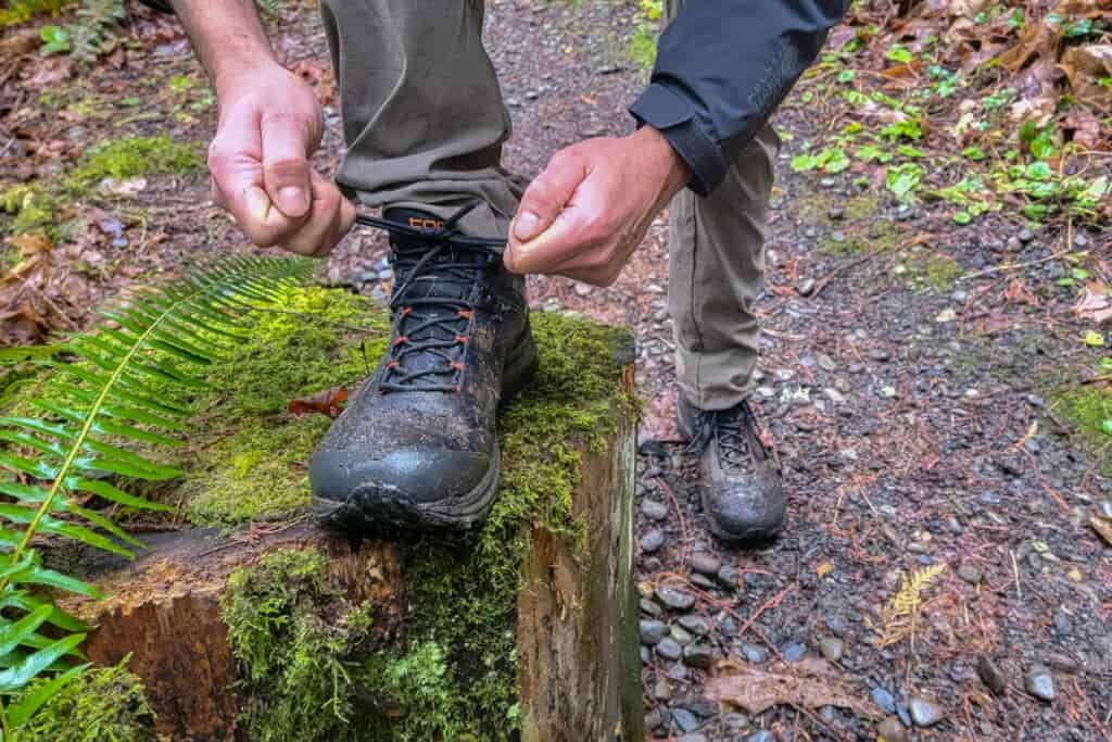 A hiker tying the laces of his muddy Topo Athletic Trailventure 2 boot up on a mossy stump