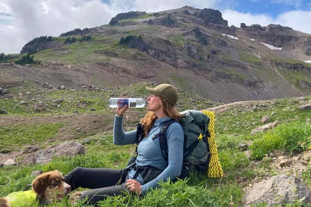 A female hikers sips water while resting next to a dog in the mountains