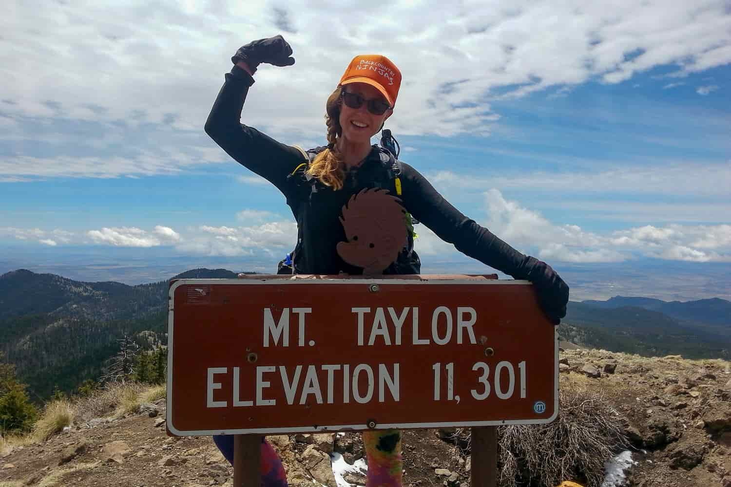 A female hiker flexes her muscles on top of a mountain behind a sign that reads Mt. Taylor Elevation 11,301