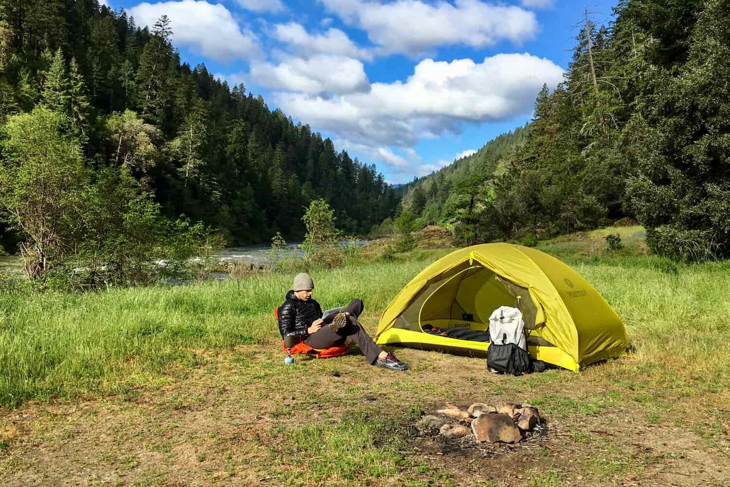 A hiker sites on a ground chair next to his yellow tent while reading