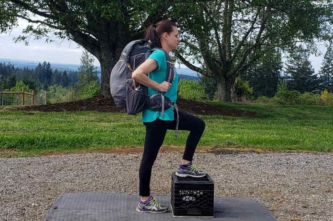 A woman steps on a milk crate on a mat while wearing a loaded backpack
